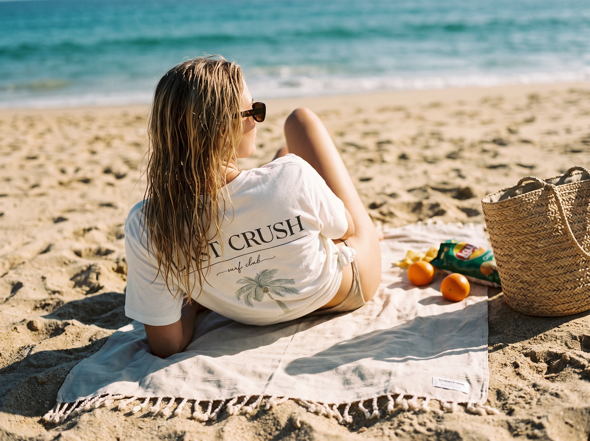 A woman sits on a beach towel on a sandy beach, viewed from behind. She is wearing the Surf Club white graphic t-shirt with 'LAST CRUSH' and a palm tree print, sunglasses, and a swimsuit.