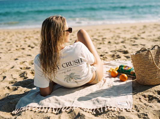 A woman sits on a beach towel on a sandy beach, viewed from behind. She is wearing the Surf Club white graphic t-shirt with 'LAST CRUSH' and a palm tree print, sunglasses, and a swimsuit.
